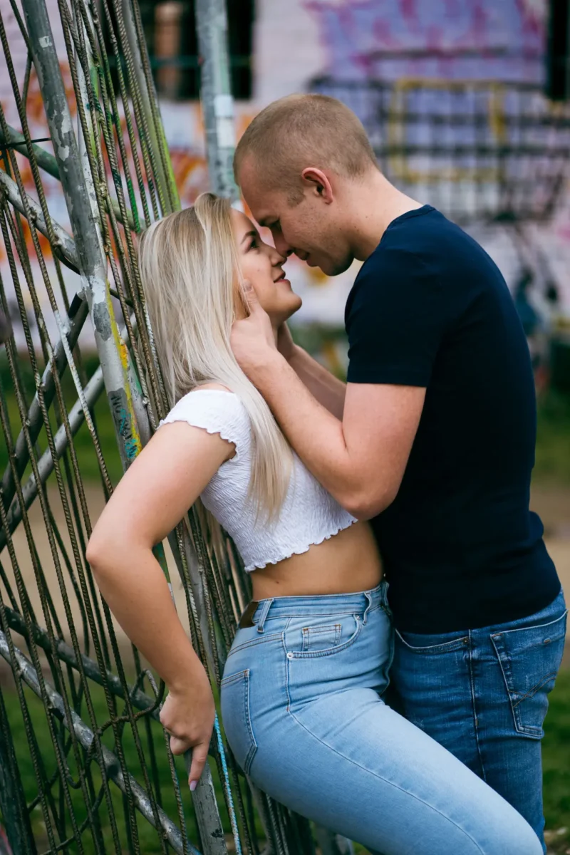 Laura und Emilis - Urbane Location in Plagwitz Paarfotograf Leipzig Plagwitz – Paar berührt sich mit Stirnen am Metallgitter, Mann fasst ihr Gesicht an