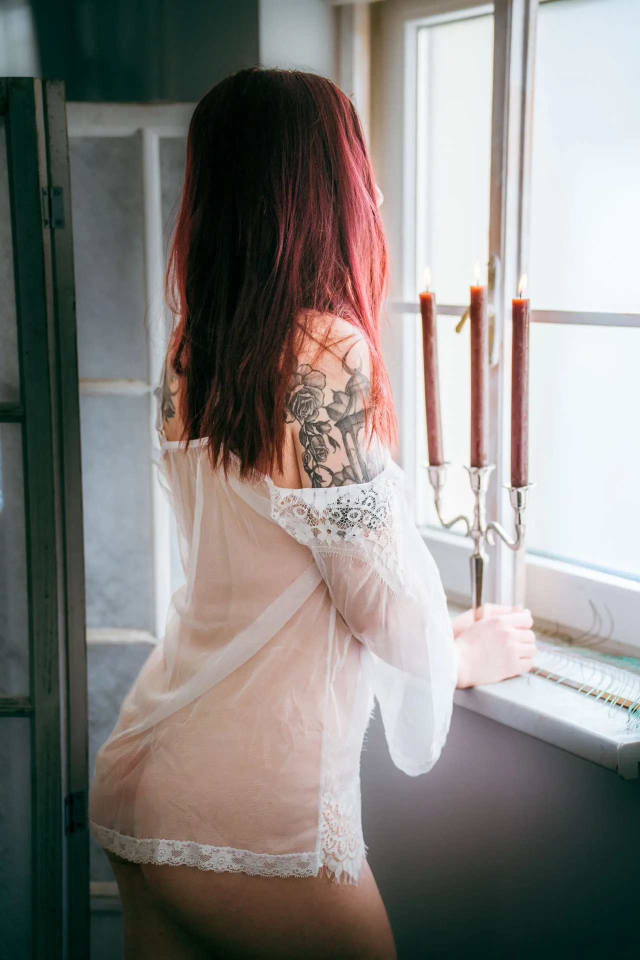 Frau im weißen transparenten Negligée steht mit dem Rücken zur Kamera am Studiofenster neben einem Kerzenständer – Boudoir Fotoshooting Leipzig