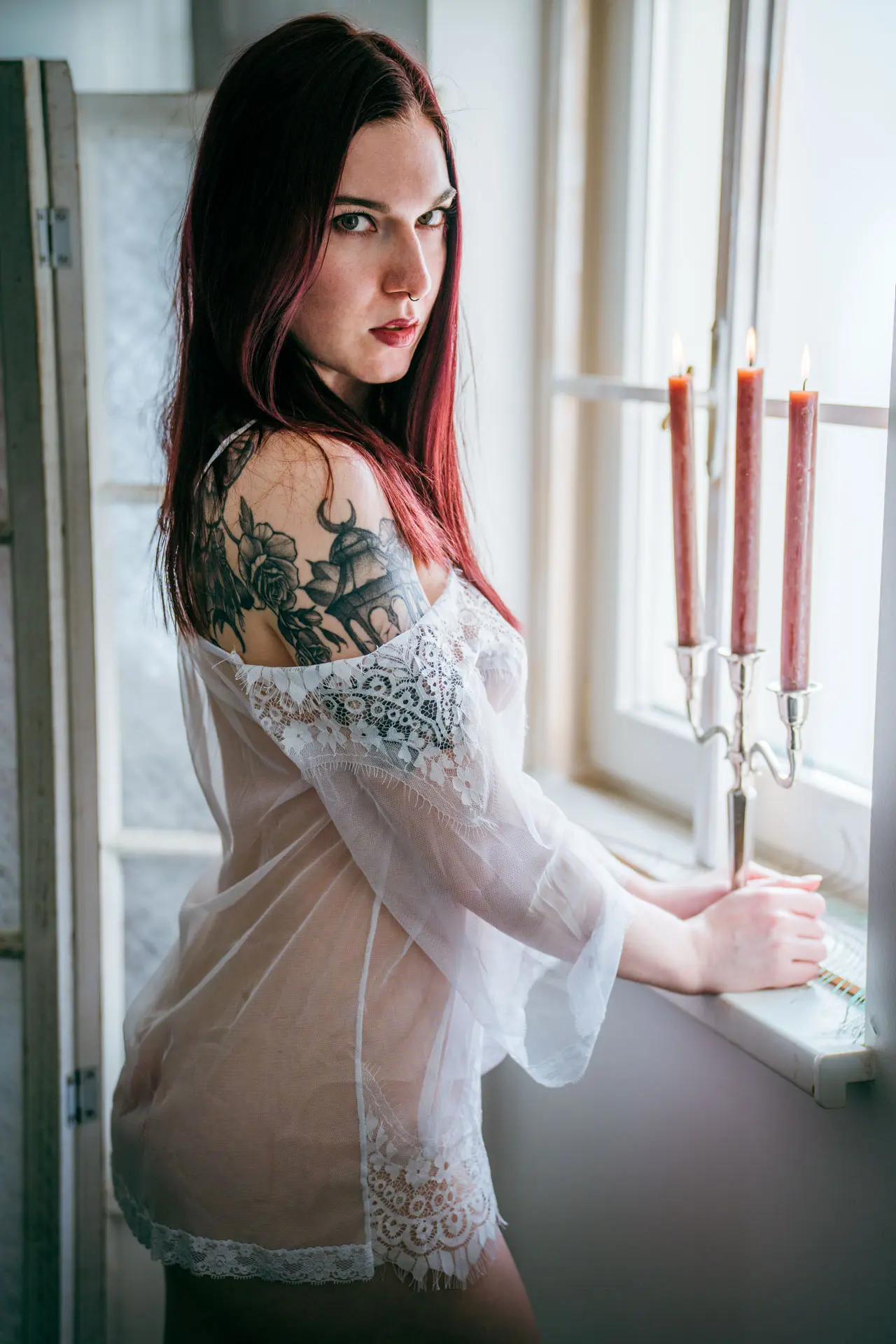 Frau im weißen transparenten Negligée dreht sich am Studiofenster zur Kamera um – intensiver Blick, Kerzenständer im Hintergrund – Boudoir Fotoshooting Leipzig