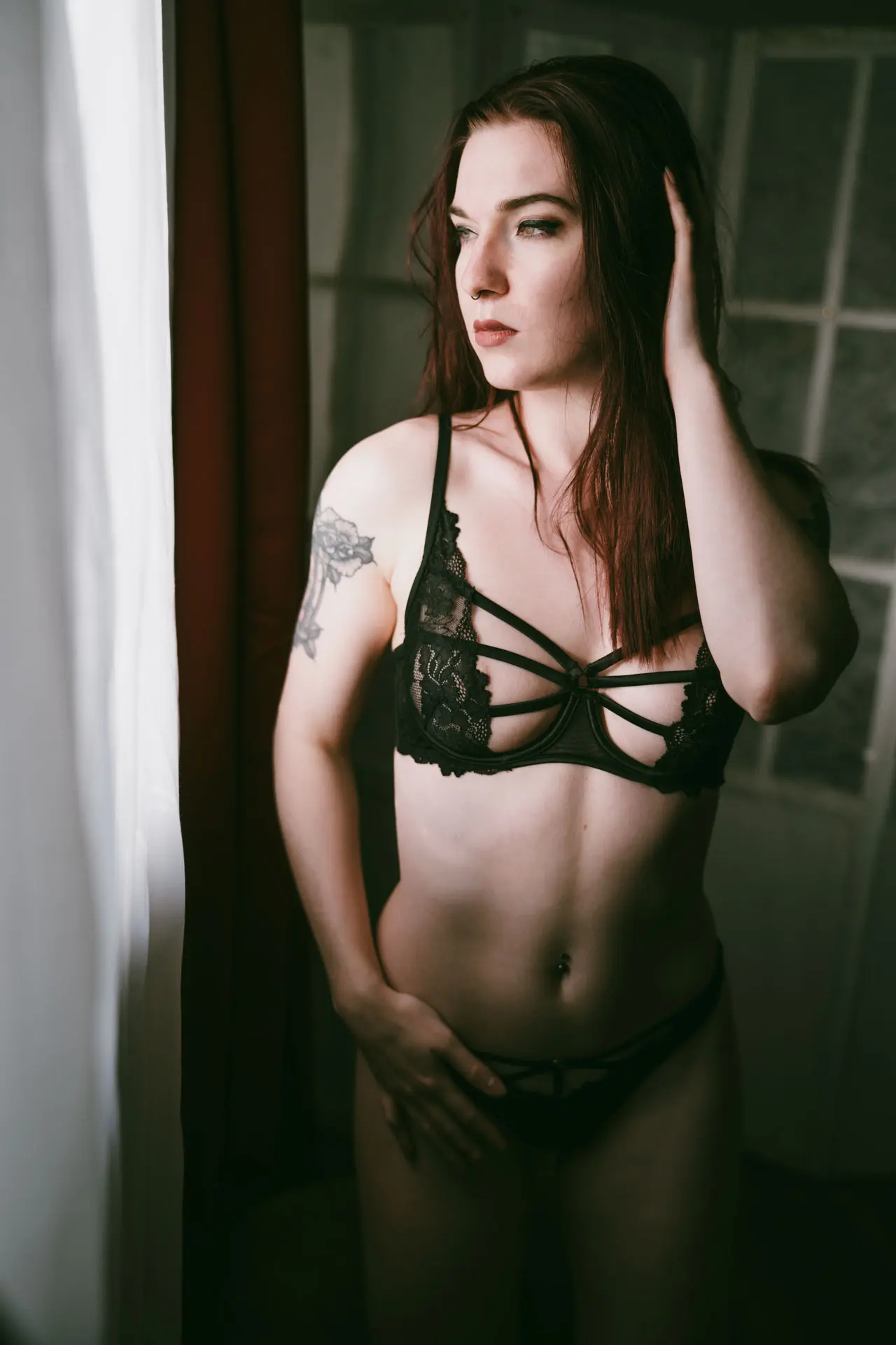 Frau in schwarzer Spitzen-Dessous steht am Studiofenster und blickt nachdenklich zur Seite – Hand in den Haaren – Boudoir Fotoshooting Leipzig