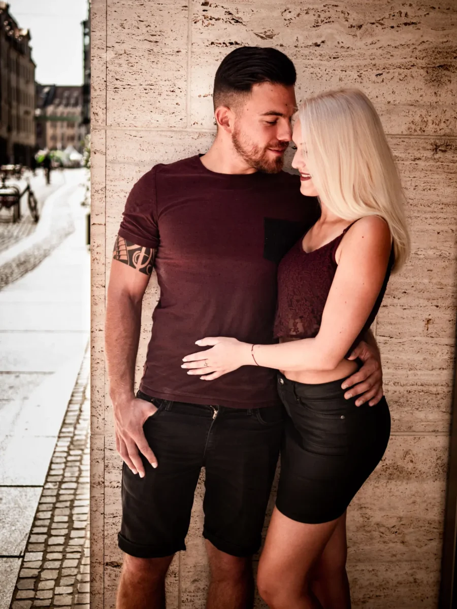 Verführerische Paarpose an einer Steinwand – blonde Frau im Lederminirock und bordeauxfarbenem Spitzentop schmiegt sich lächelnd an ihren Partner im engen Shirt